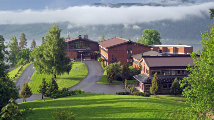 Oversiktsbilde over Honne. Hotellet og skogkurs lokaler fra utsiden. Honne ligger på toppen av en bakke, med mjøsa og fjell på andre siden i bakgrunnen.