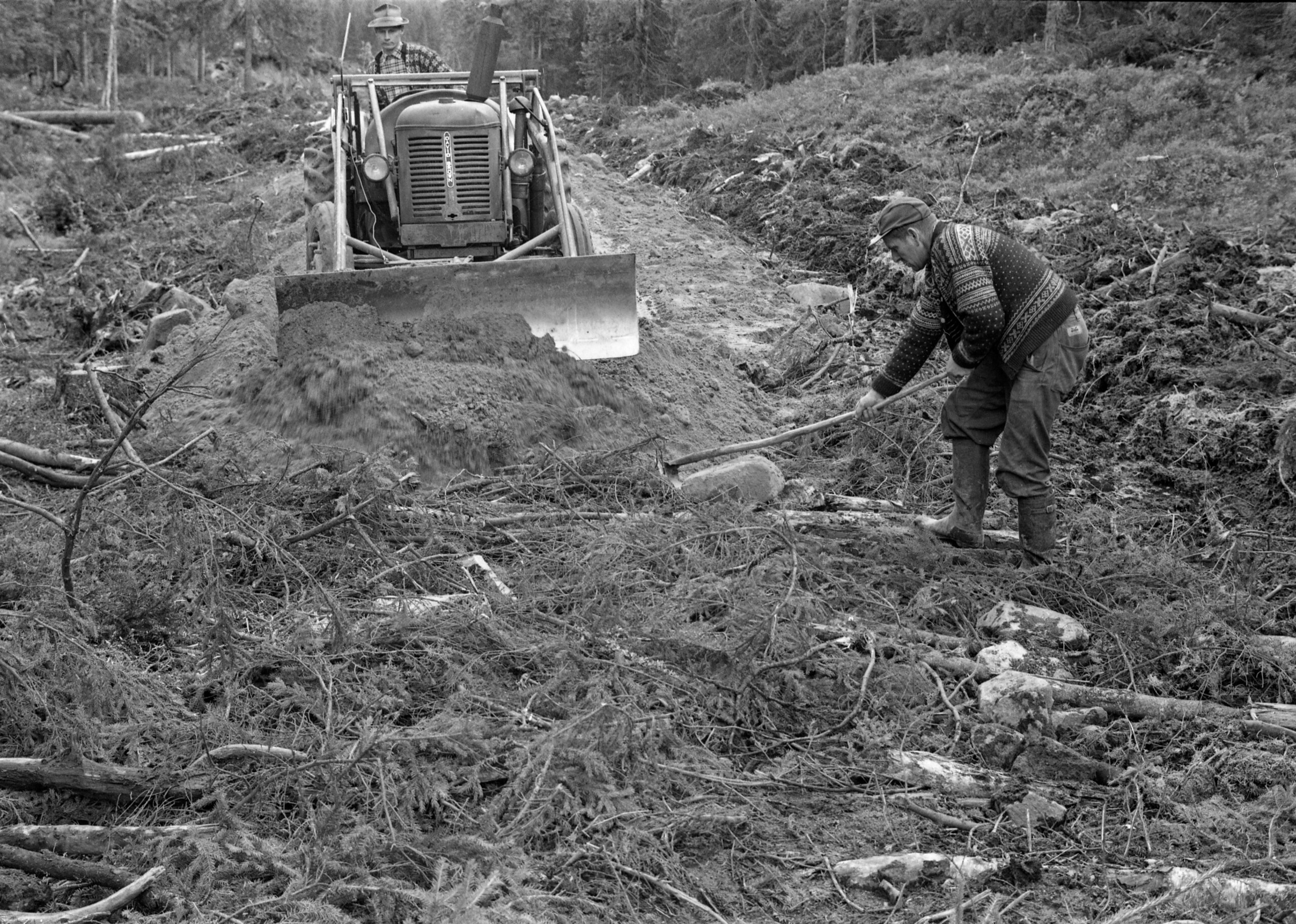 Vegbygging på fuktig mark i Treschow Fritzøes skoger i 1964. Her ser vi to menn og en traktor i aktivitet ved det som på dette tidspunktet var fronten i vegprosjektet. Fotografiet viser hvordan man hadde lagt hogstavfall – småtrær og kvist – i det som skulle bli veglinja, for å gi bedre bæring for de massene vegen skulle bygges av. Disse massene ble skjøvet på plass ved hjelp av en David Brown-traktor med frontmontert skjær med bistand fra en mann med krafse.
