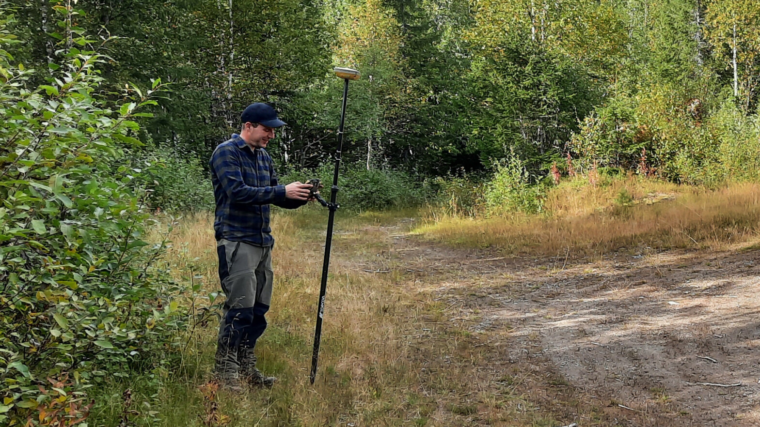 Mann som såt på en gjenngrodd vei. Han har en GPS mottaker på en stang og gjør innmålinger. Foto. nger